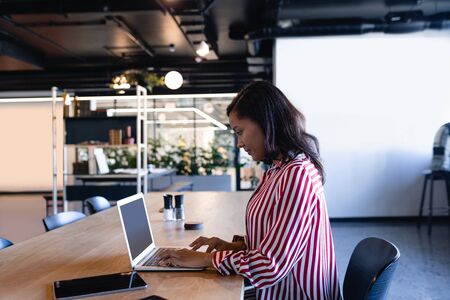 Side view of a mixed race businesswoman working in a modern office, sitting by a desk and using a laptop computer. Social distancing and self isolation in quarantine lockdownの写真素材