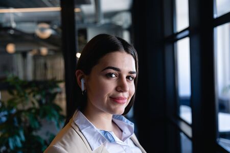 Side view close up of a Caucasian woman wearing smart clothes and earphones, working in the modern office, standing by the window and looking straight into a camera. Social distancing and self isolation in quarantine lockdownの写真素材
