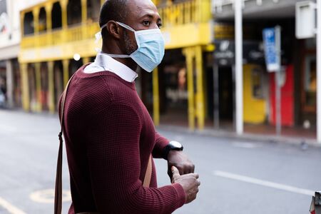 African American man out and about in the city streets during the day, wearing a face mask against air pollution and covid19 coronavirus, crossing the street and using his smartwatch.の写真素材