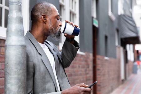 African American man out and about in the city streets during the day, standing on the street, drinking a takeaway coffee and using his smartphone.の写真素材