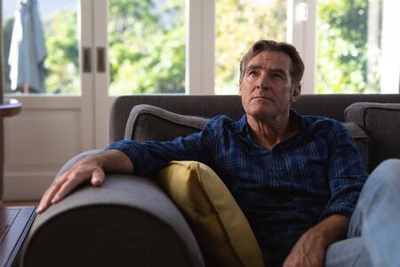Senior Caucasian man enjoying his time at home, sitting on a couch, looking away from the camera,...