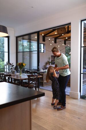 Caucasian man enjoying his time at home, dancing with his daughter in a kitchen, smiling, social distancing and self isolation in quarantine lockdown during coronavirus covid19 epidemicの写真素材