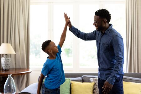 African American man enjoying his time at home, standing by a couch, embracing his son, high fiving...