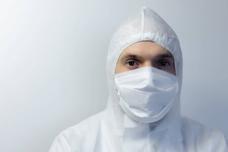 Portrait of a Caucasian man wearing lab overalls and a face mask standing in an empty white room,...