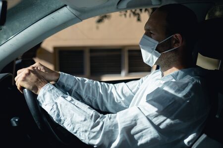 Caucasian man sitting in a car and driving, wearing a face mask against covid 19 and a white shirt on a sunny dayの写真素材