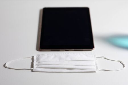 Close up of a tablet and white face mask protection against pandemic coronvirus covid 19 and for safety lying on a white tableの写真素材