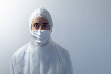 Portrait of a Caucasian woman wearing lab overalls and a face mask standing in an empty white room,...