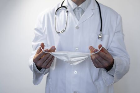 Caucasian man, wearing doctor uniform and a stethoscope on his shoulders, wearing a face mask and standing in an empty white roomの写真素材
