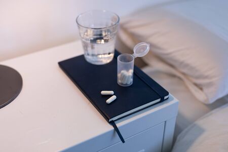 Close up of a notebook, box of tablets and a glass of water lying on a cabinet next to a bed,...