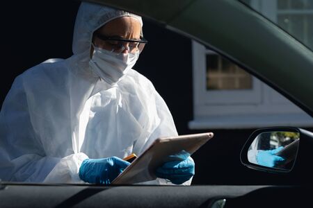 Caucasian woman, wearing safety uniform, wearing a face mask and standing outside by a car, using...