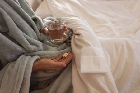 Mid section of sick Caucasian woman spending time at home self isolating and social distancing in quarantine lockdown during coronavirus covid 19 epidemic, sitting up in bed taking tabletsの写真素材