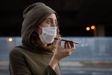 Caucasian woman out and about in the city streets during the day, wearing a face mask against covid19 coronavirus talking on her smartphoneの写真素材