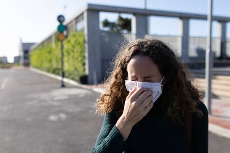 Caucasian woman out and about in the city streets during the day, wearing a face mask against covid19 coronavirus covering her face while sneezingの写真素材