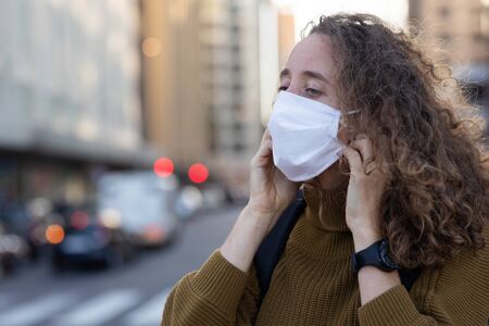 Caucasian woman out and about in the city streets during the day, putting on a face mask against...