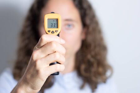 Portrait of a Caucasian woman with long curly hair, examining her temperature, holding a non contact gun thermometer, social distancing and self isolating during coronavirus outbreak.の写真素材