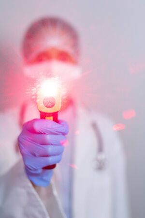Portrait of a Caucasian female healthcare worker wearing a lab coat, face mask and gloves against coronavirus, covid 19, holding a non contact thermometer and looking straight into a camera.の写真素材