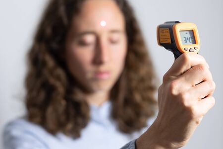 Caucasian woman with long curly hair, examining her temperature, holding a non contact gun...