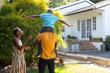 African American woman spending time with her partner who is giving their son a piggyback in the garden. Social distancing and self isolation in quarantine lockdown.の写真素材