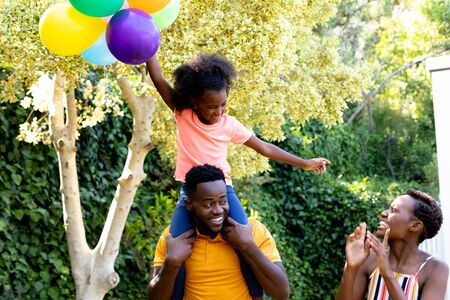 African American woman spending time with her partner who is giving their daughter a piggyback in the garden. Social distancing and self isolation in quarantine lockdown.の写真素材