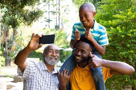 Senior African American man spending time with his son and his grandson in the garden on a sunny day, taking a selfie with a smartphone.の写真素材