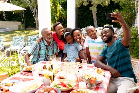 African American family spending time together in the garden on a sunny day, sitting by a table and having a lunch, while a man is taking a selfie with his smartphone.の写真素材