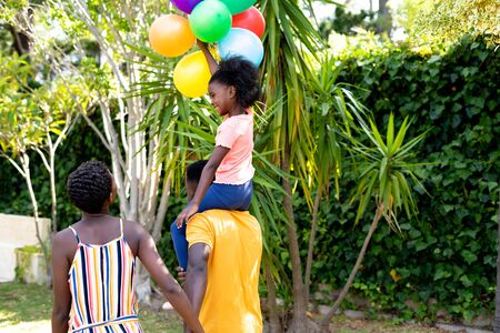 Rear view of an African American woman spending time with her partner who is giving their daughter a piggyback in the garden. Social distancing and self isolation in quarantine lockdown.の写真素材