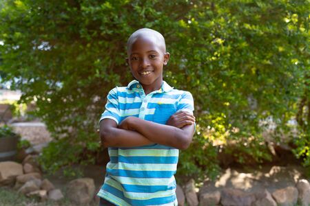 Portrait of an African American boy wearing stripes polo shirt, spending time in the garden, standing with his arms crossed and smiling. Social distancing and self isolation in quarantine lockdown.の写真素材