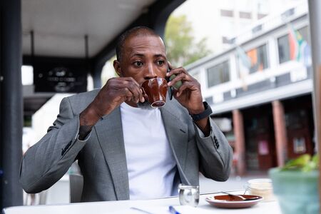 African American man spending time at cafe, sitting at a coffee terrace, having a cup of coffee and talking on his smartphone.の写真素材