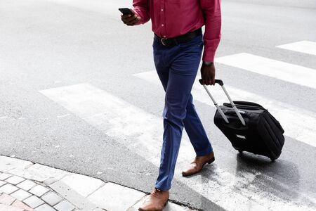 Low section of African American man out and about in the city streets during the day, wearing pink shirt crossing the street, using a smartphone and pulling a suitcase.の写真素材