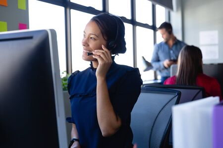 Front view of a mixed race woman wearing headset, working in a modern office, using a desktop...