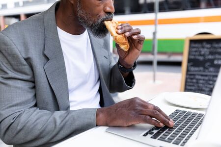 African American man spending time at cafe, sitting at a coffee terrace, having a snack and using a laptop computer.の写真素材