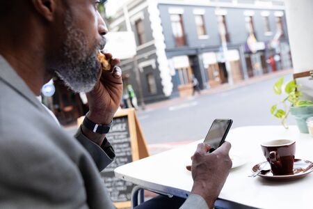African American man spending time at cafe, sitting at a coffee terrace, having a snack and using his smartphone.の写真素材
