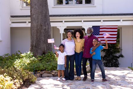 Multi-generation mixed race family enjoying their time at a garden, welcoming a visitor, embracing, kids are holding mini flags, with big flag in the backgroundの写真素材