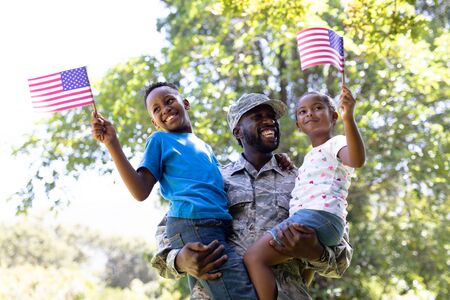 African American man wearing a military uniform, returning home, standing in a garden, holding and embracing his children, the kids are holding and waving mini flagsの写真素材