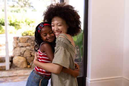 Mixed race girl enjoying her time at home, standing in a hallway, greeting her mother, embracing and smiling, with front doors openの写真素材