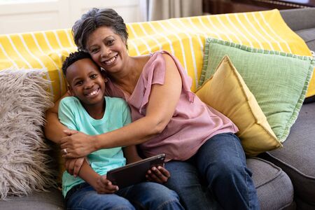 Mixed race boy and his grandmother enjoying their time at home together, sitting on a couch, using a...