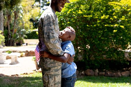 African American male solider wearing uniform embracing his son holding a USA flag, standing in...