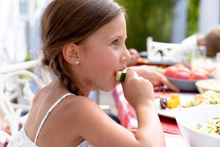Caucasian girl sitting by a table in a garden on a sunny day and eating a slice of watermelon during a family lunch.の写真素材