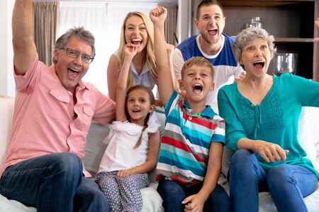 Three generation Caucasian family spending time together at home, sitting on a couch and watching television, having fun and cheering.の写真素材