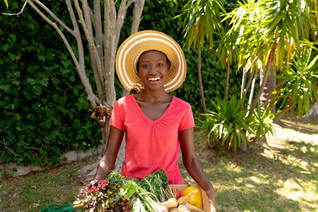 African American woman spending time in the, garden smiling and presenting fresh vegetables. Social distancing and self isolation in quarantine lockdown.の写真素材