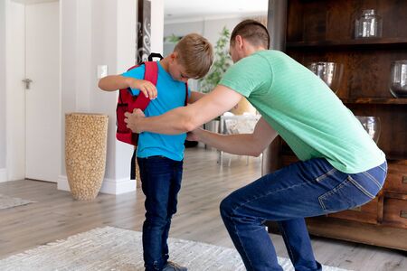 Caucasian man wearing a green t-shirt helping his son wearing a red rucksack preparing for leaving...