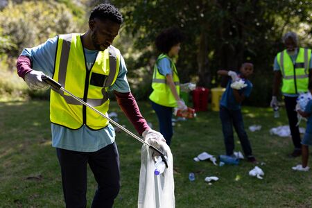 Multi-generation mixed race family spending time outside together, all wearing blue volunteer t shirts and protective gloves, collecting garbage, holding garbage bags, on a sunny dayの写真素材