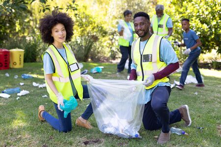 Portrait of a mixed race couple spending time outside with their family, presenting a garbage bag with garbage in it, looking at the camera and smiling, with their family in the background, all wearing blue volunteers t shirts, on a sunny dayの写真素材
