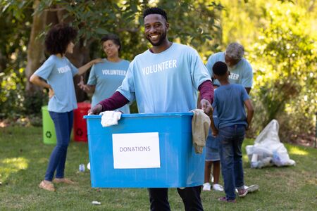 Portrait of an African American man spending time outside with his family, presenting a donations box with clothes in it, looking at the camera and smiling, with his family in the background, all wearing blue volunteers t shirts, on a sunny dayの写真素材