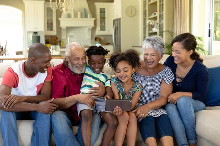 Front view of a multi-generation mixed race family at home in the living room, sitting on a couch together and looking at a tablet computer that the granddaughter is holdingの写真素材