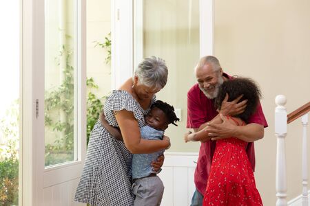 Front view of a mixed race senior couple arriving for a visit, smiling and embracing their young grandson and granddaughter in the hallway of their homeの写真素材