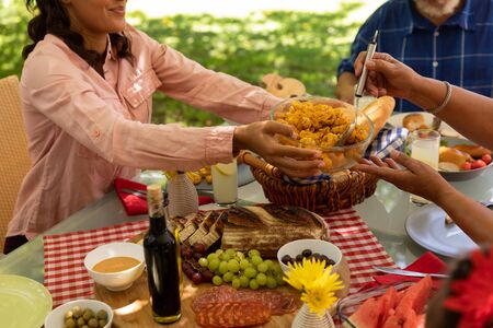 Side view mid section of a mixed race woman holding a dish white her senior mother serves herself food, her father in the background, sitting at a table for a family meal together outside on a patio in the sunの写真素材