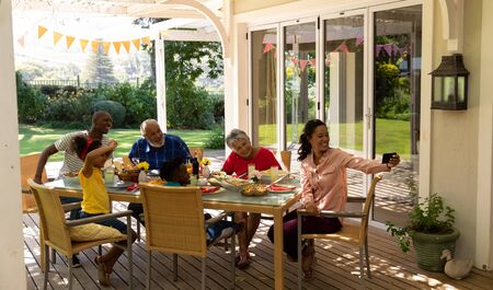 Side view of a multi-ethnic, multi-generation family sitting at a table for a meal together outside on a patio in the sun, the mixed race mother using a smartphone to take a selfie with them all smilingの写真素材