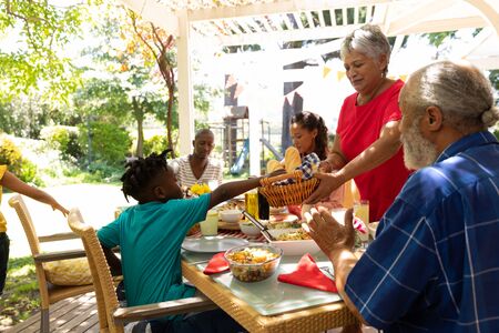 Side view of a multi-ethnic, multi-generation family serving food and sitting down at a table for a...