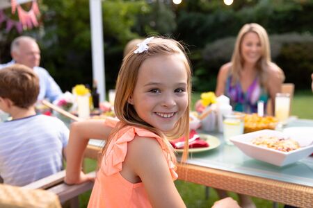 Portrait of a Caucasian girl sitting at a table and turning to camera smiling during a meal outside in the garden with her multi-generation familyの写真素材